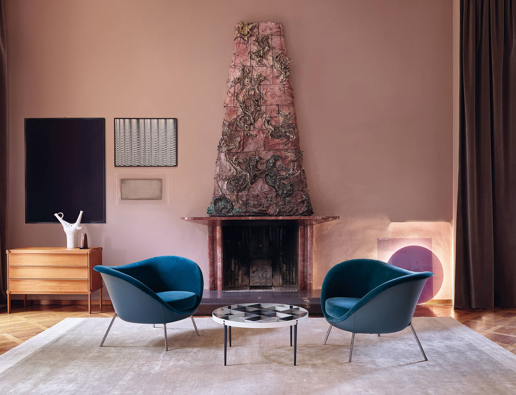 Living room with two teal velvet armchairs, a round coffee table and a tall decorative ceramic fireplace surround