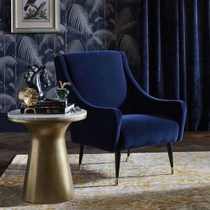 Navy blue velvet armchair with brass legs beside a round brass side table against botanical wallpaper