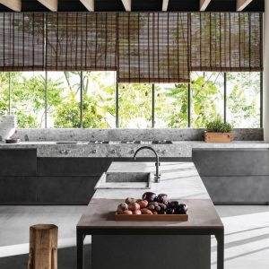 Concrete kitchen island with a stone countertop, bowl of fruit and bamboo blinds by a leafy window