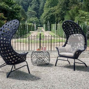 Two black wicker high-backed lounge chairs and a round side table on gravel facing a formal garden